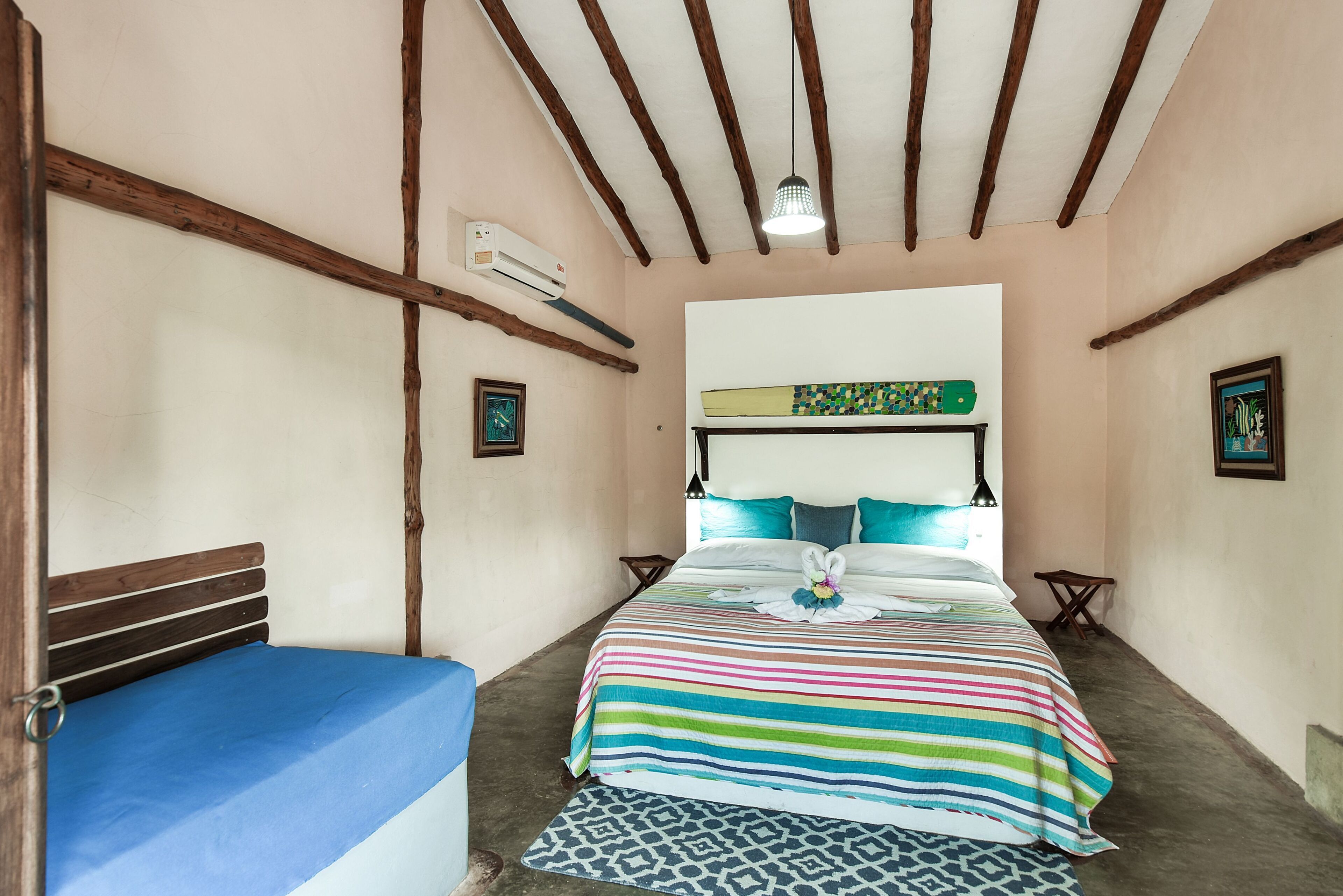 Un dormitorio con cama, colchón azul, colcha a rayas coloridas, cabecera, vigas de madera en el techo, unidad de aire acondicionado y una pequeña mesa con una silla.
