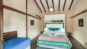 Un dormitorio con cama, colchón azul, colcha a rayas coloridas, cabecera, vigas de madera en el techo, unidad de aire acondicionado y una pequeña mesa con una silla.
