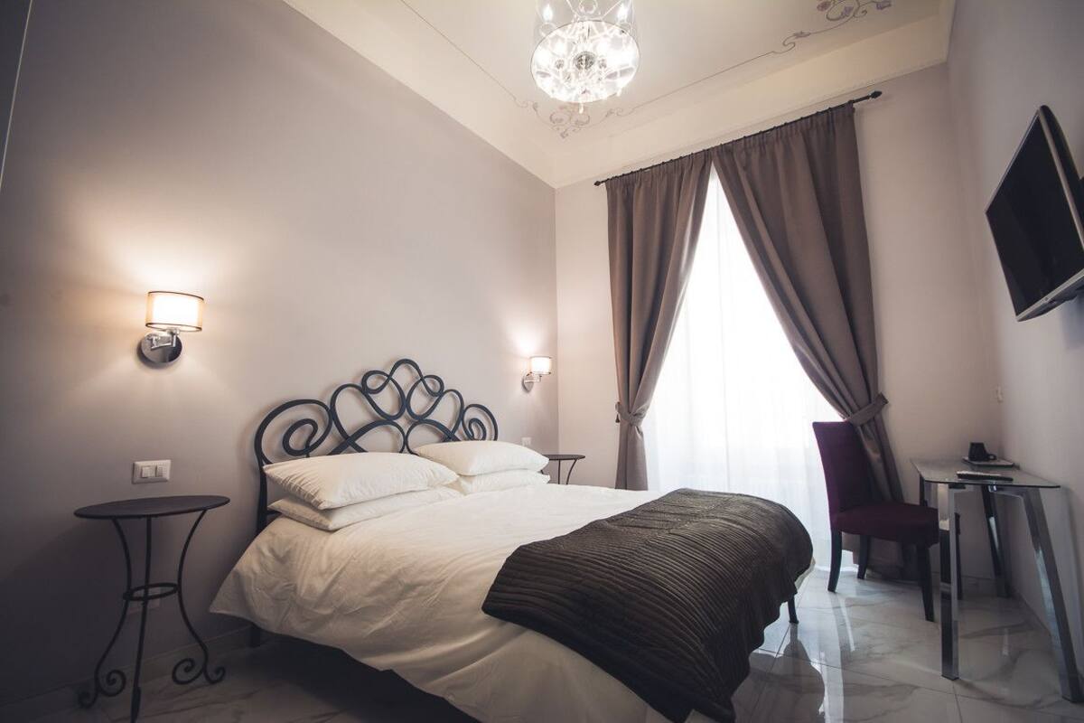 A hotel room with a bed, a desk, a chair, a TV, and a chandelier.