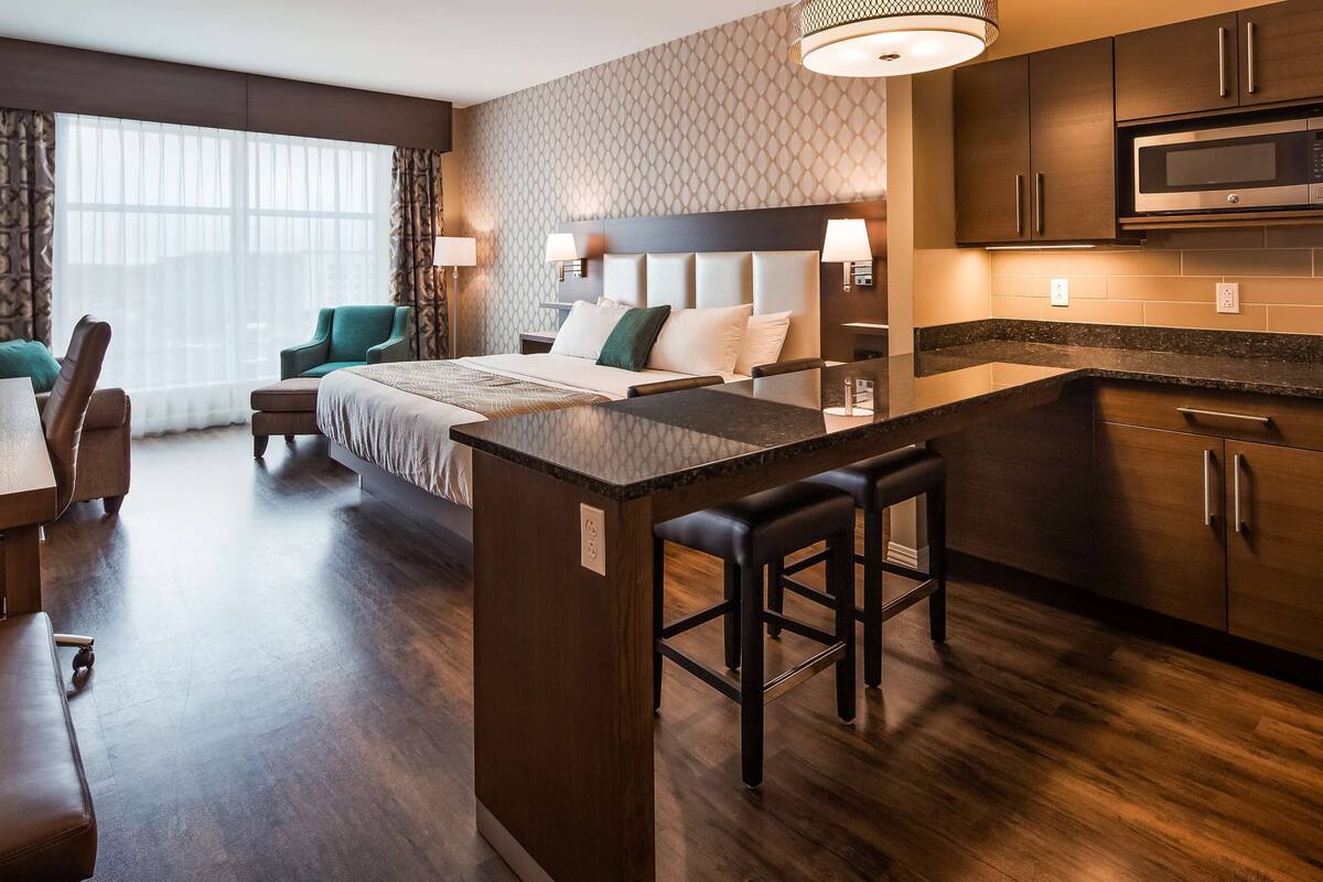 A modern hotel room with a kitchenette, a dining area, and a large bed with a headboard.