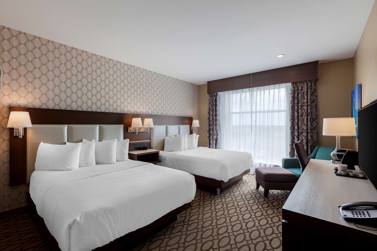 A hotel room with two beds, a desk, a chair, a TV, and a large window with curtains.