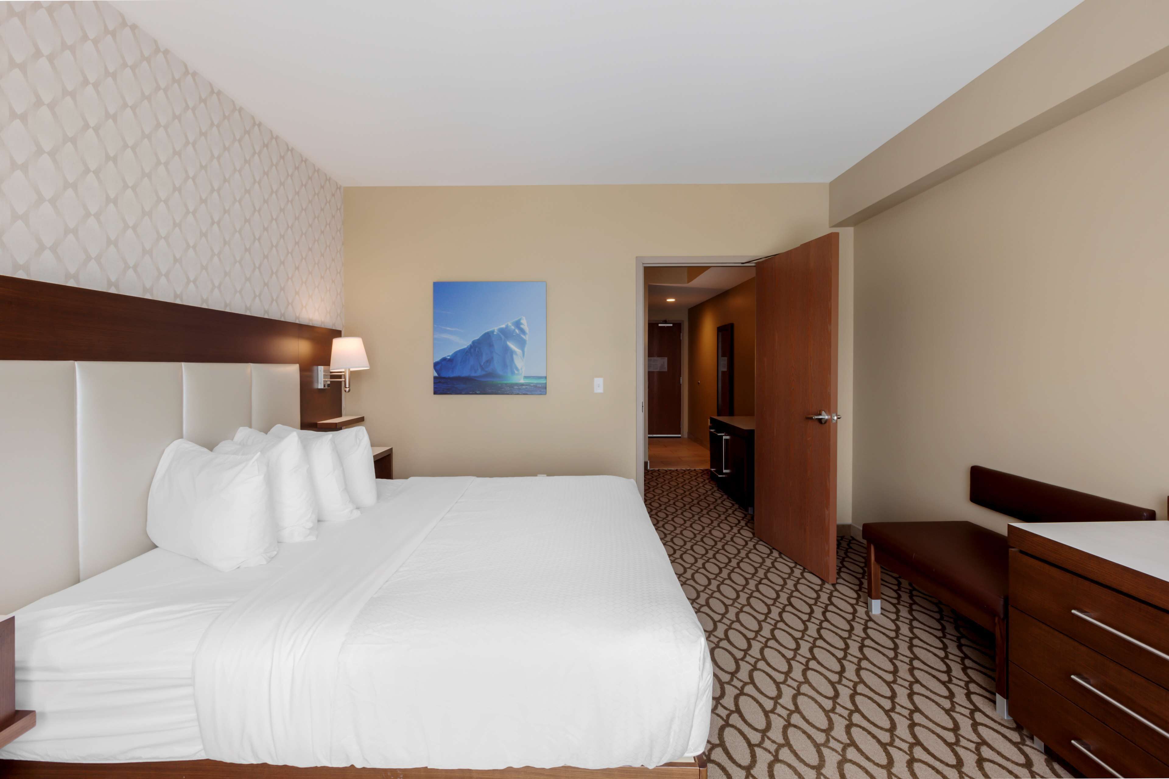 Ein Hotelzimmer mit einem großen Bett, einem Nachttisch mit Lampe, einem Holzkopfschild, einem an der Wand hängenden Bild von einem Eisberg, einem gemusterten Teppich und einer Holzbank.