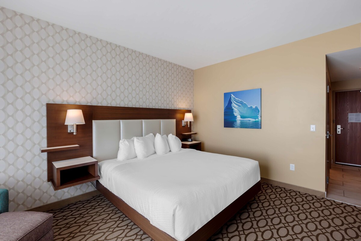 A hotel room with a large bed, a headboard with built-in lighting, a nightstand, a wall-mounted shelf, and a painting of a mountain landscape.