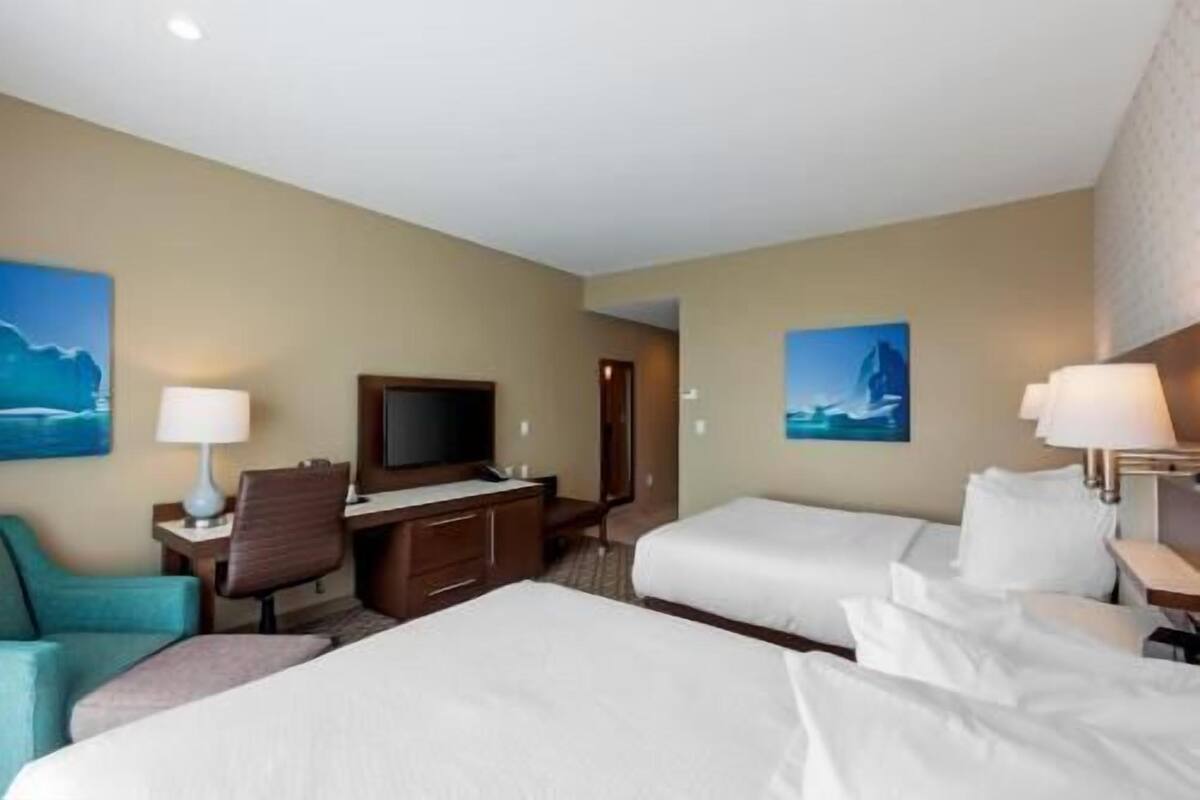 A hotel room with two beds, a desk with a chair, a TV, and two paintings on the walls.