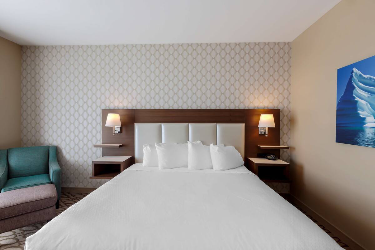 A hotel room with a large bed, two bedside tables with lamps, a teal armchair, and a patterned wallpaper.