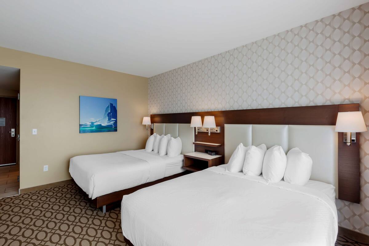 A hotel room with two beds, a wooden headboard, a nightstand, and a wall-mounted lamp.