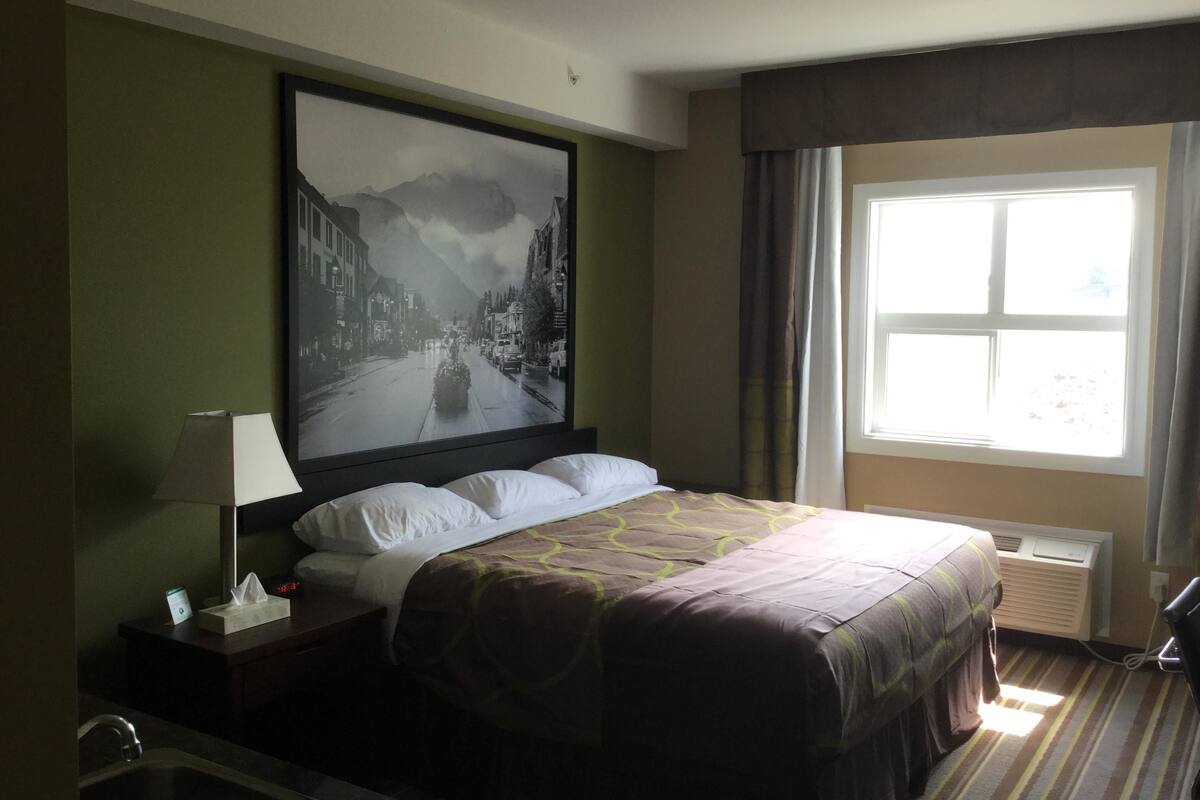 Habitación de hotel con una cama, una mesita de noche con lámpara, un cuadro enmarcado en la pared y una ventana con cortinas.