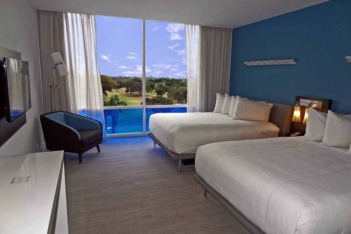 A hotel room with two beds, a desk, a chair, and a large window with a view of greenery.