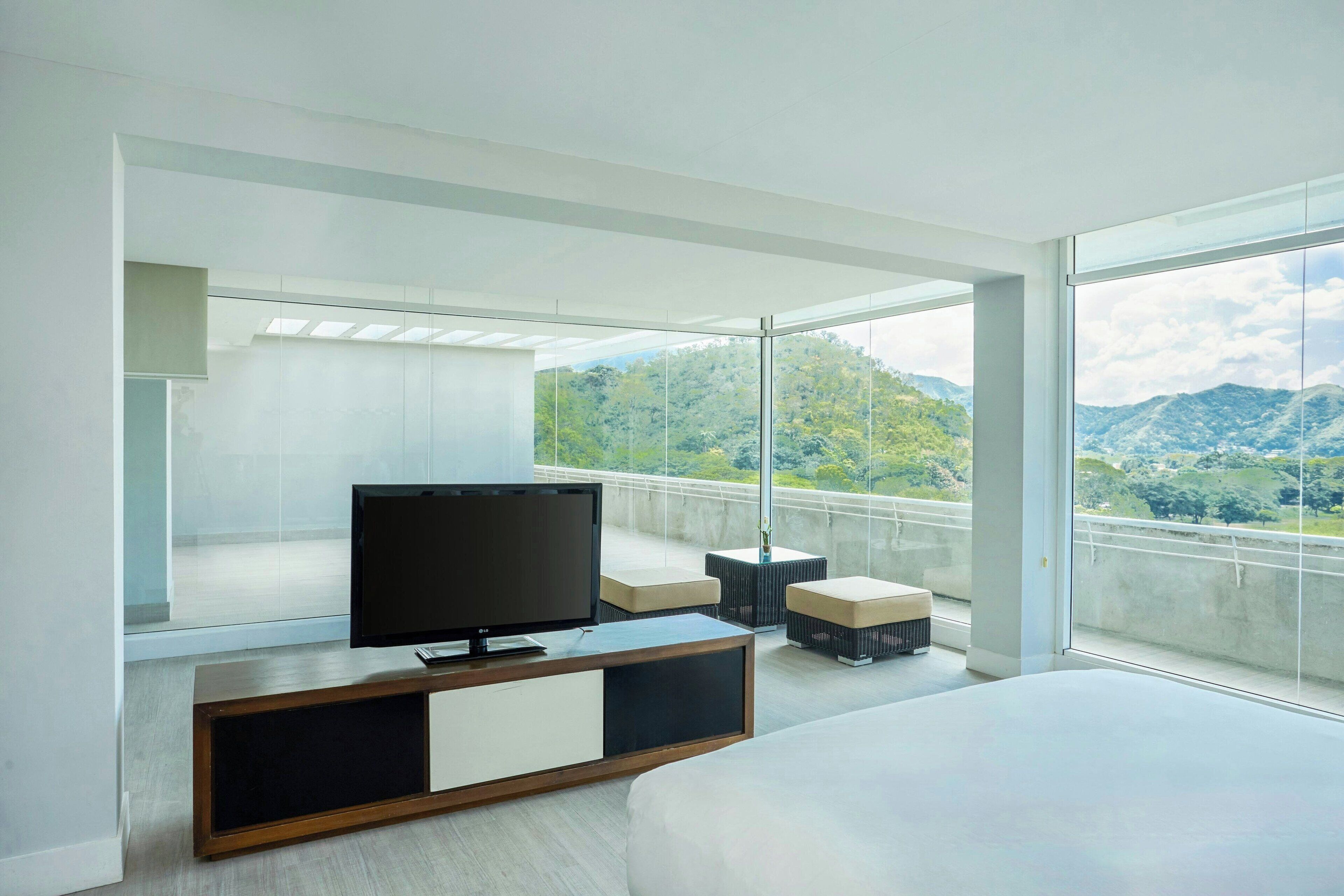 A modern hotel room with a large bed, a TV on a wooden console, and a balcony with a mountain view.