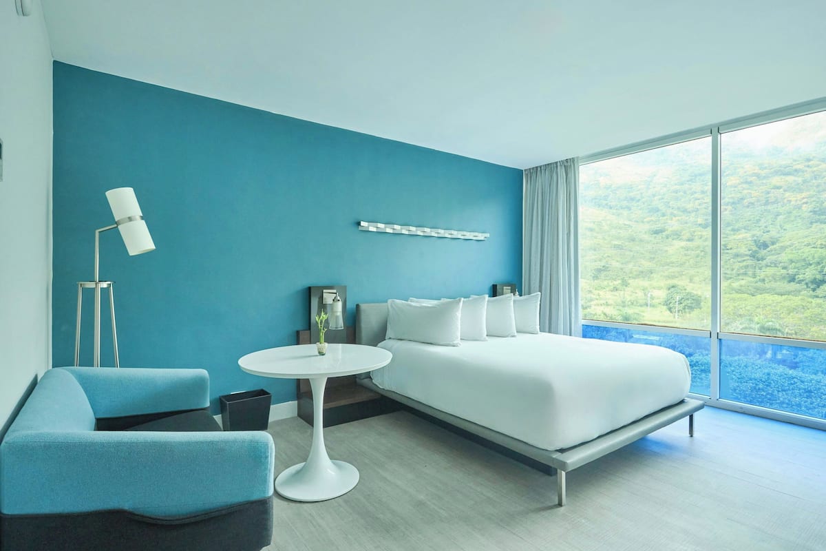 A modern hotel room with a large bed, a blue accent wall, a round white table, and a view of a lush landscape through large windows.