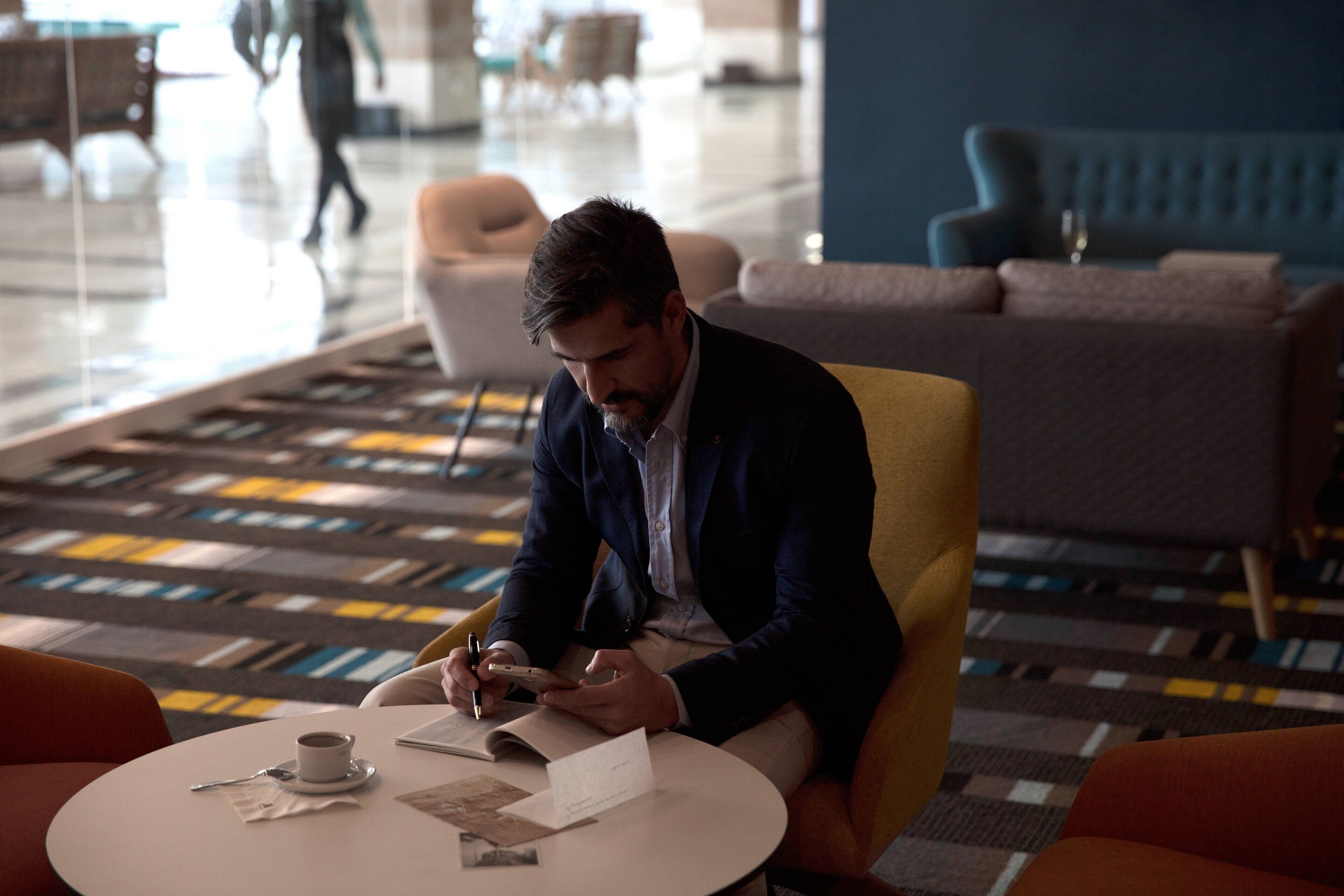 Um homem sentado a uma mesa redonda numa área de estar moderna com um tapete estampado, uma chávena de café e alguns documentos.