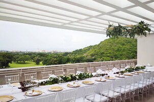 An outdoor dining area with a long table set for a meal, surrounded by white chairs, and overlooking a lush green landscape with a city in the distance.