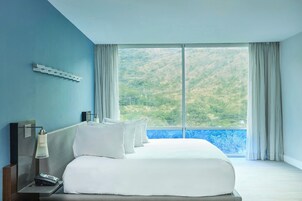 A hotel room with a large bed, a nightstand with a lamp, and a view of a mountainous landscape through a large window.