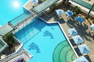 An aerial view of a swimming pool with dolphin murals, surrounded by umbrellas and palm trees.