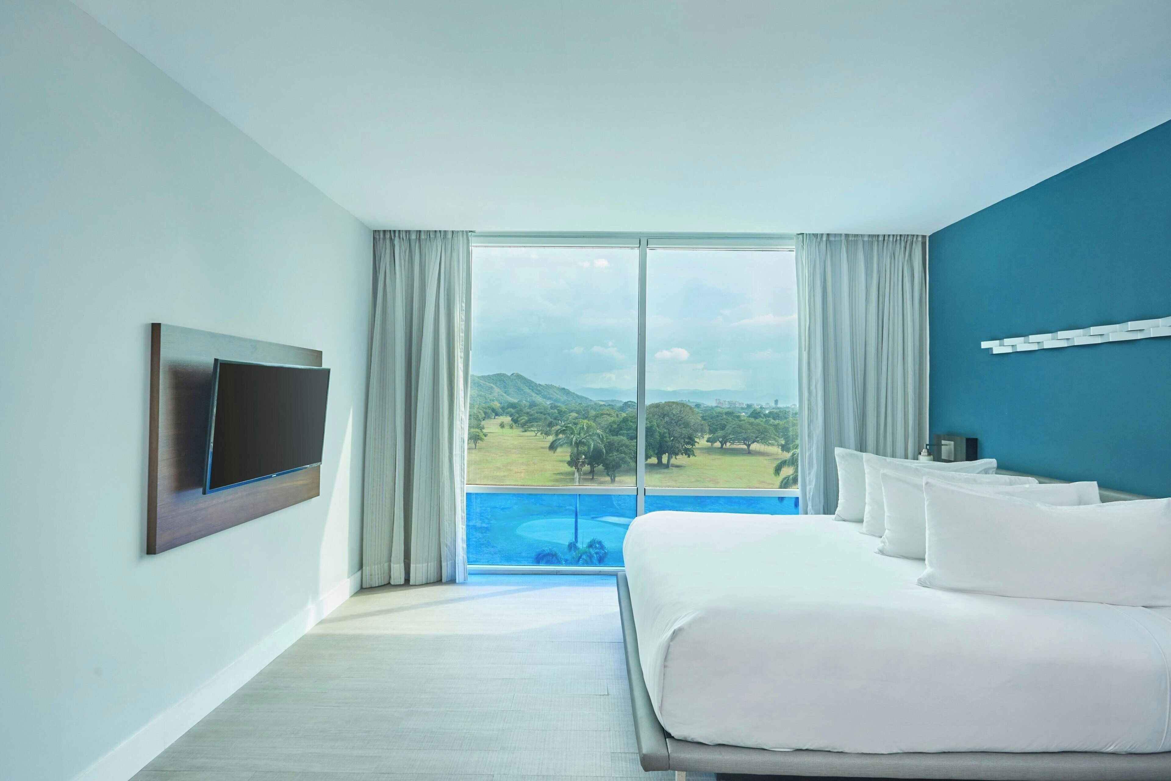 Habitación de hotel con una cama grande, un televisor de pantalla plana montado en la pared y vistas a un campo de golf y montañas a través de la ventana.