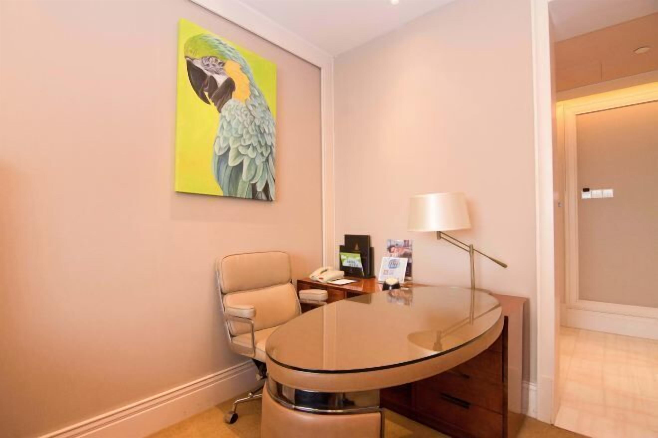 A hotel room with a glass-top desk, a chair, a lamp, a telephone, and a framed picture on the wall.
