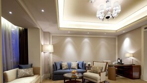 A well-lit living room with a sofa, armchair, and coffee table, featuring a chandelier and a desk with a lamp. - Juva Grand Hotel (Hohhot)