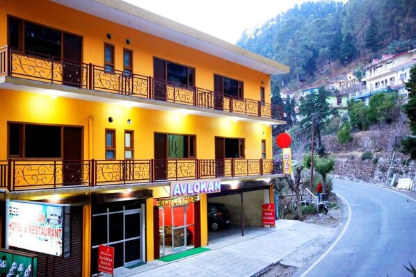 Un hôtel à deux étages doté de balcons, d’une enseigne indiquant « Avlokan Hotel & Restaurant », et offrant une vue sur un paysage vallonné.