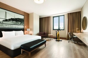 A hotel room with a large bed, a desk, a chair, a small table, a treadmill, and a large window with a city view.
