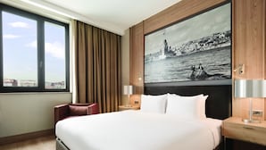 Habitación de hotel con una cama grande, vista de un paisaje urbano a través de una ventana, un cuadro de una barca en un río y una mesita de noche de madera con una lámpara.