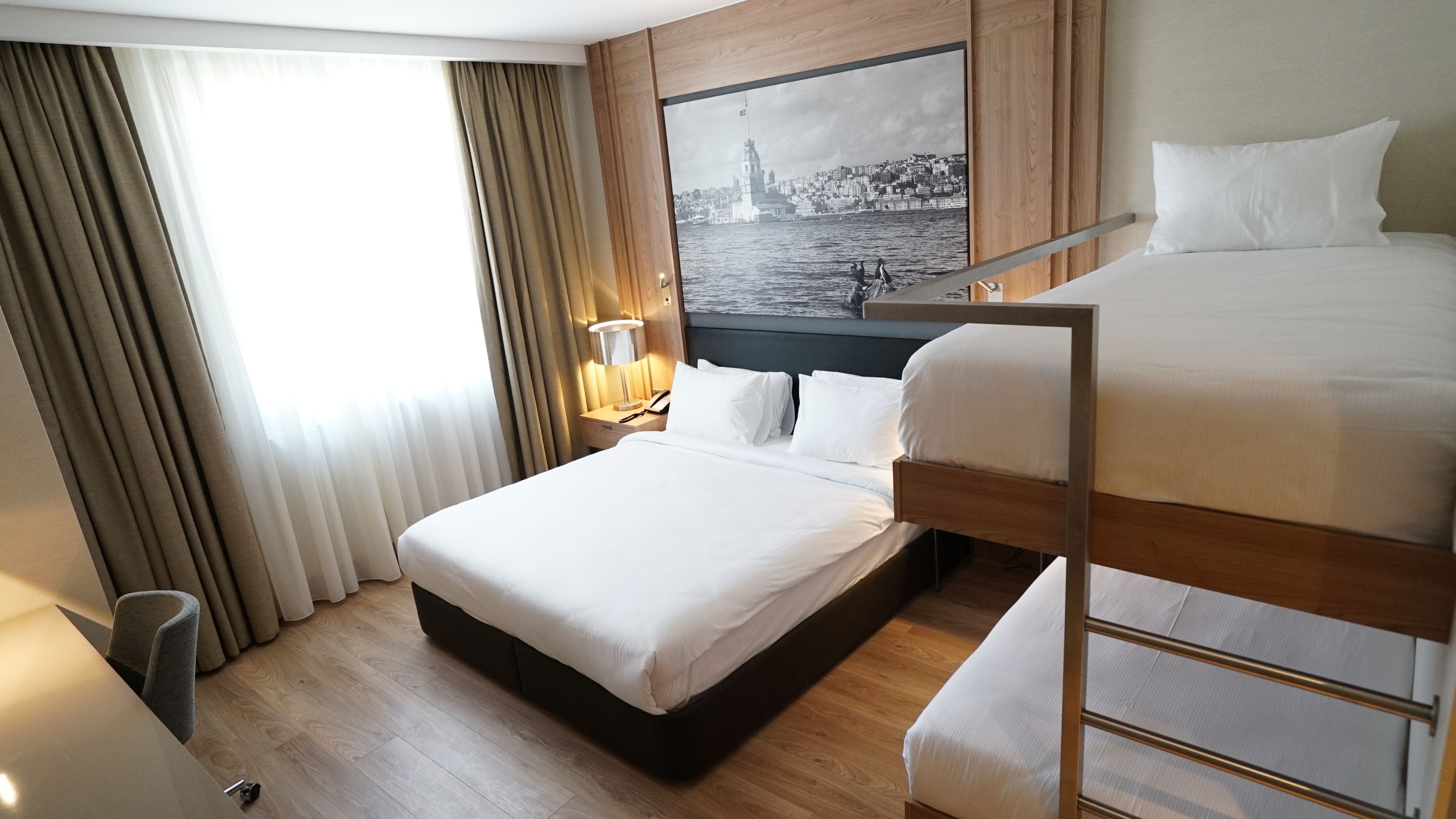 A hotel room with a large bed, a smaller bed, a nightstand with a lamp, a wooden headboard, and a cityscape painting on the wall.