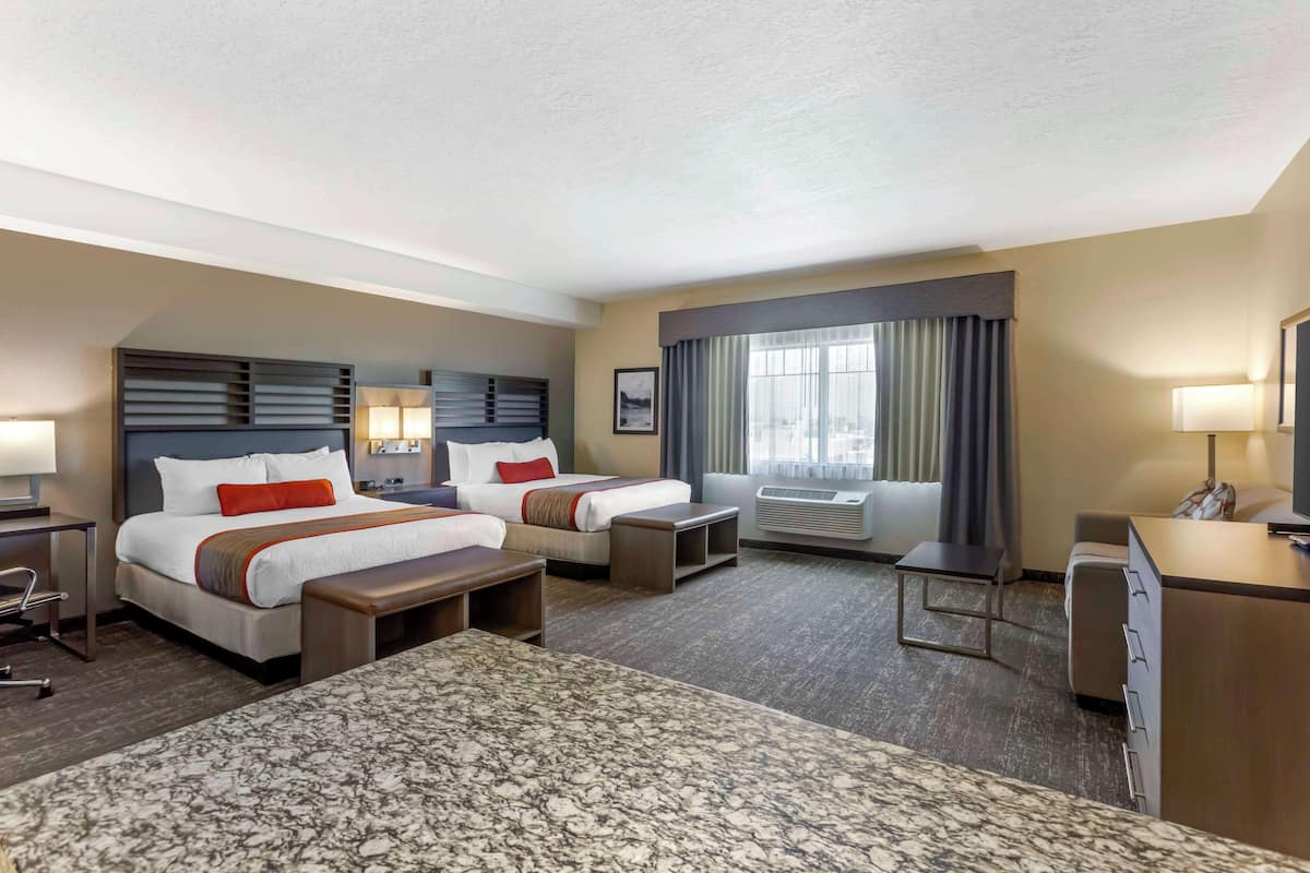 A hotel room with two beds, a desk, a chair, a small table, a sofa, and a large window with curtains.
