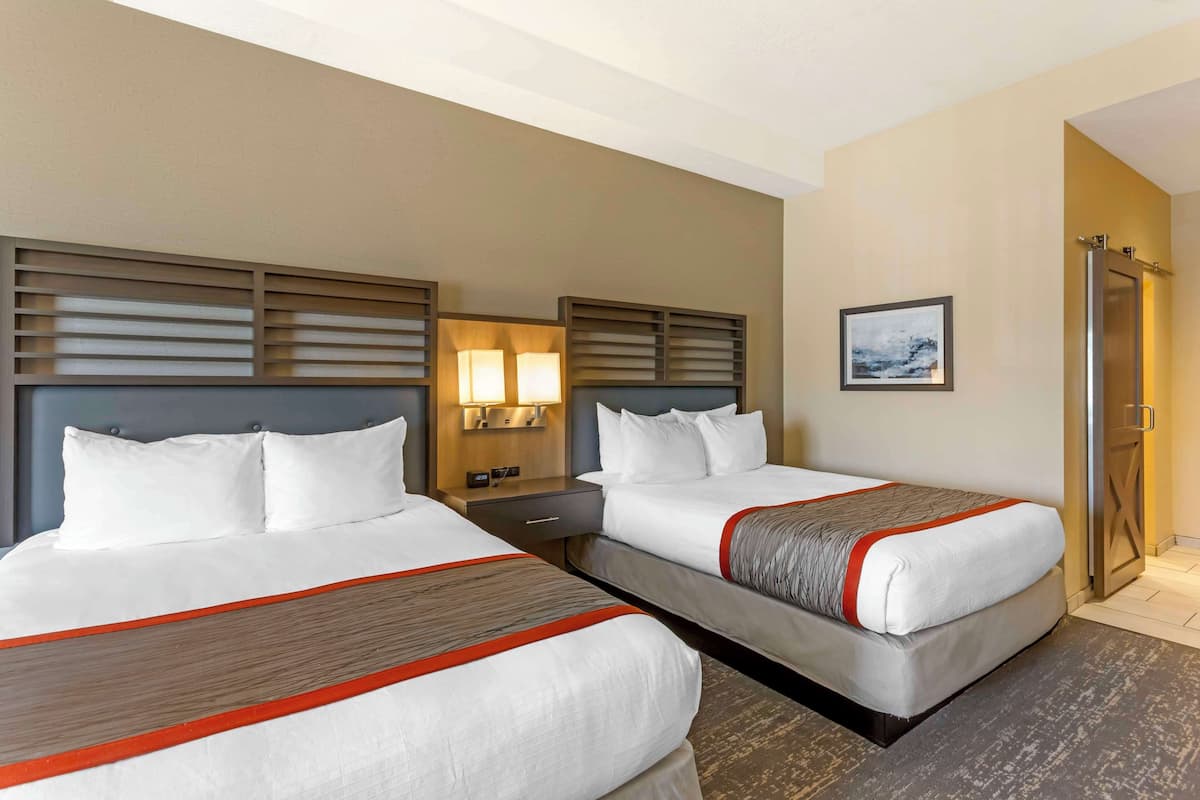 A hotel room with two beds, each with a wooden headboard, white bedding, and a red accent. There is a nightstand with a lamp between the beds, and a framed picture on the wall.
