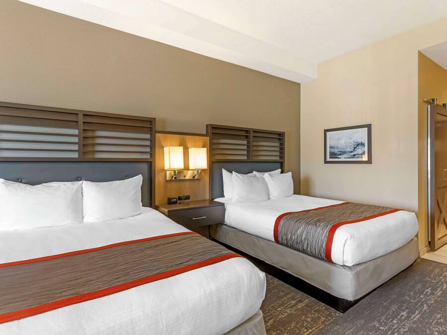 A hotel room with two beds, each with a wooden headboard, white bedding, and a red accent. There is a nightstand with a lamp between the beds, and a framed picture on the wall.