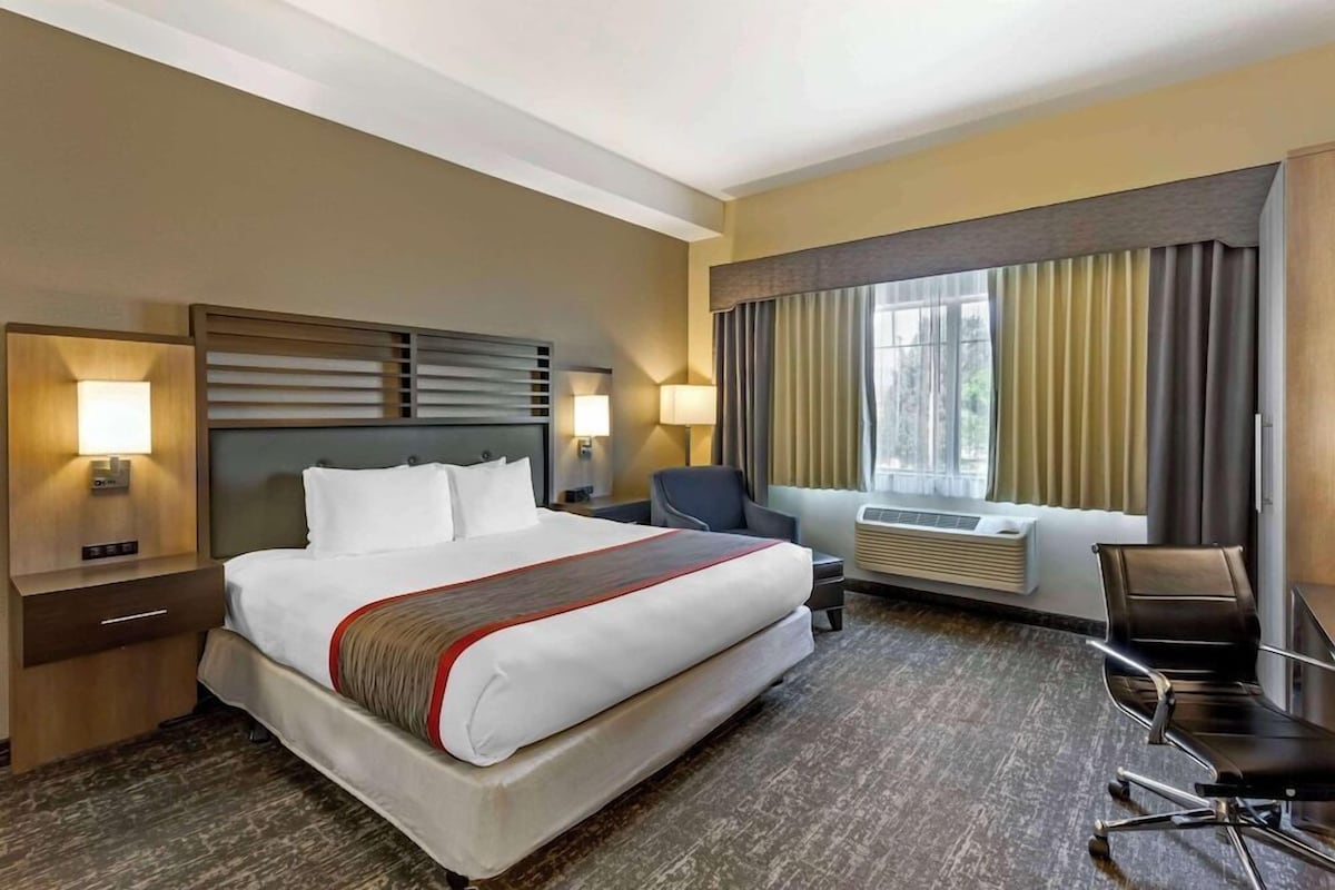 A hotel room with a large bed, bedside tables, a desk, a chair, a window with curtains, and a wall-mounted air conditioning unit.