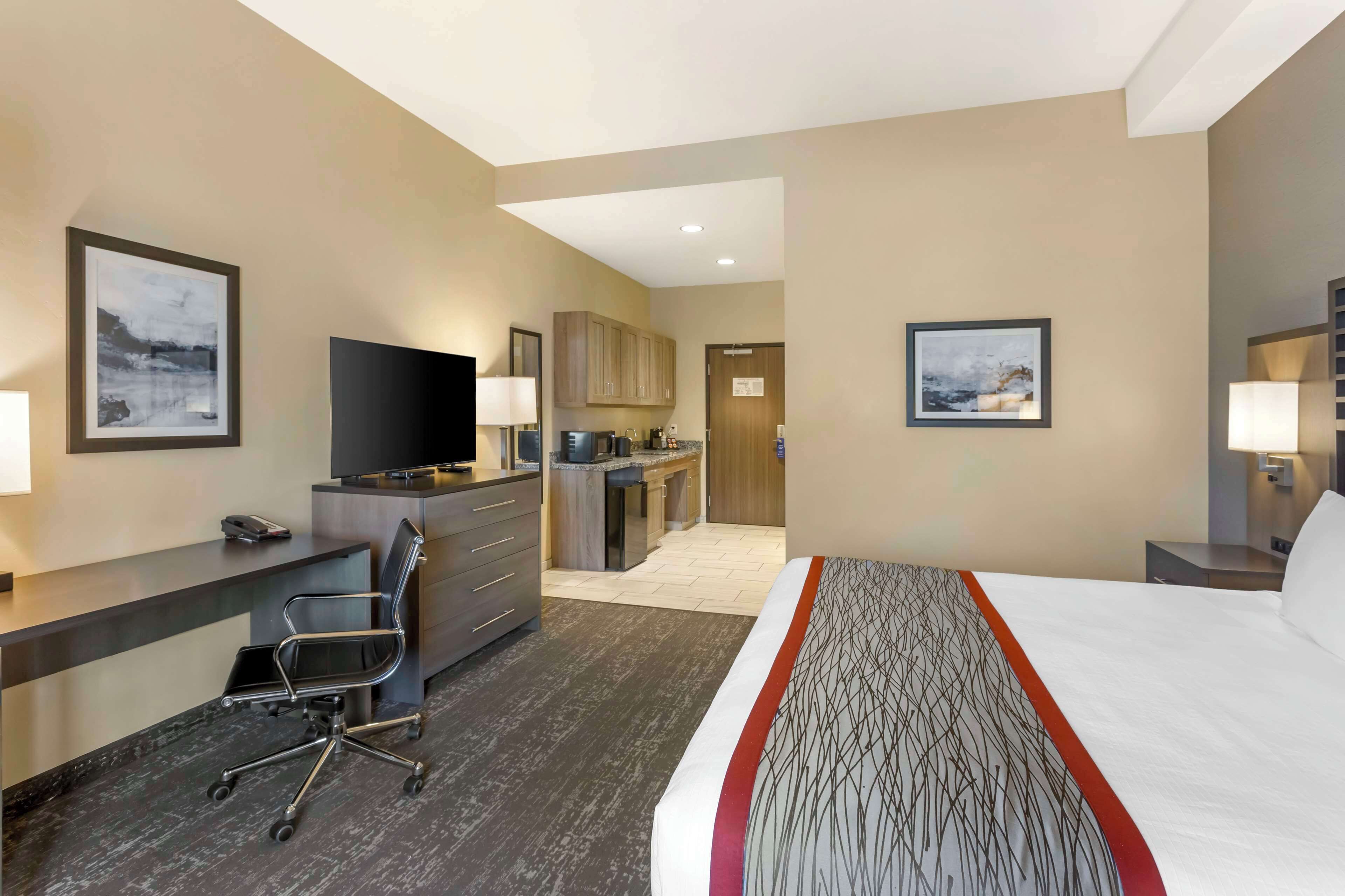 A hotel room with a bed, desk, chair, television, kitchenette, and artwork on the walls.
