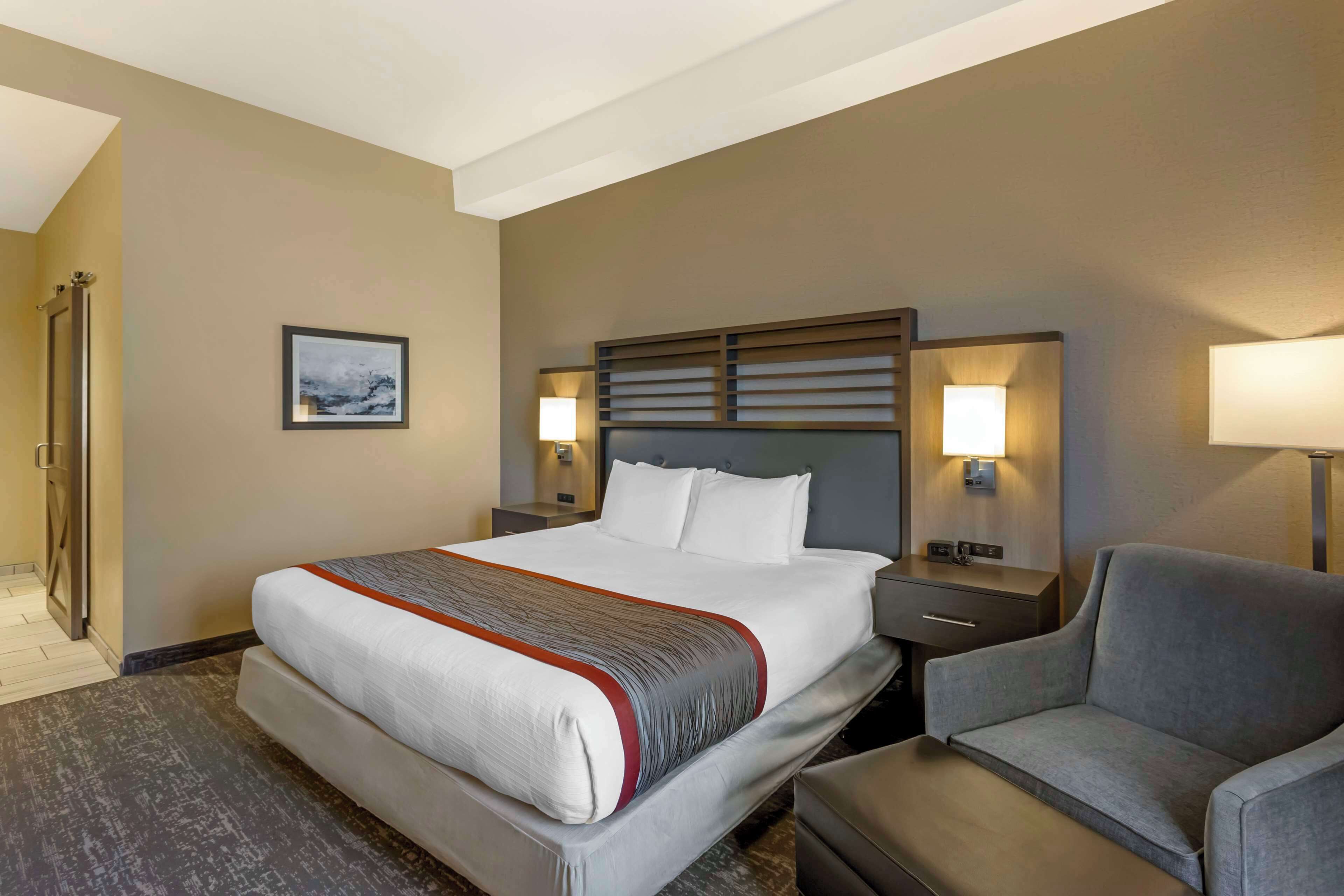 A hotel room with a large bed, two bedside lamps, a gray armchair, and a small table.