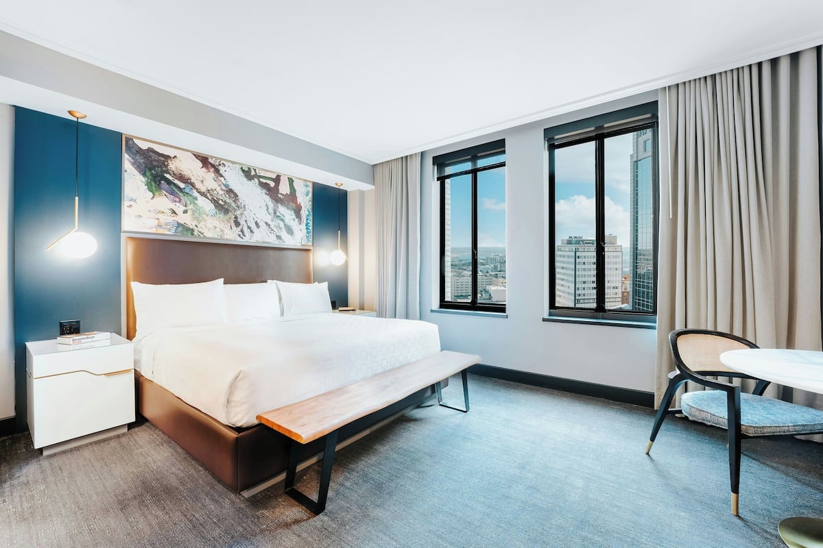 A modern hotel room with a large bed, a desk, and a chair. There is a city view through the window.