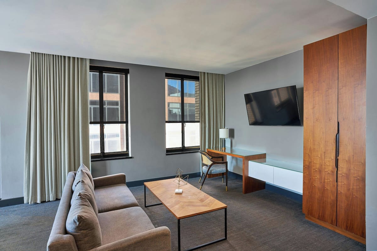 A modern hotel room with a sofa, a wooden coffee table, a desk with a chair, a television, and large windows with curtains.