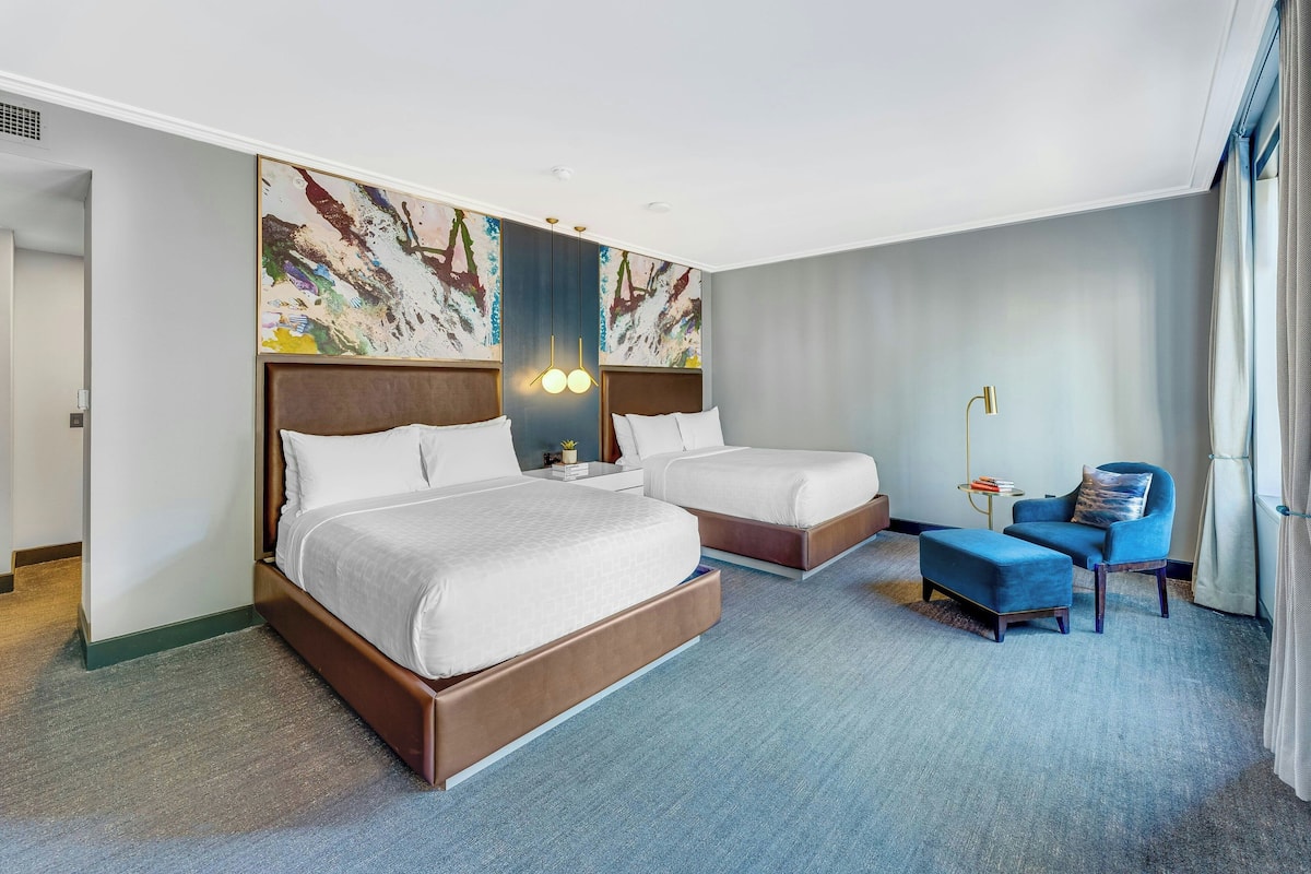 A hotel room with two beds, a blue armchair, a small table, and a painting on the wall.