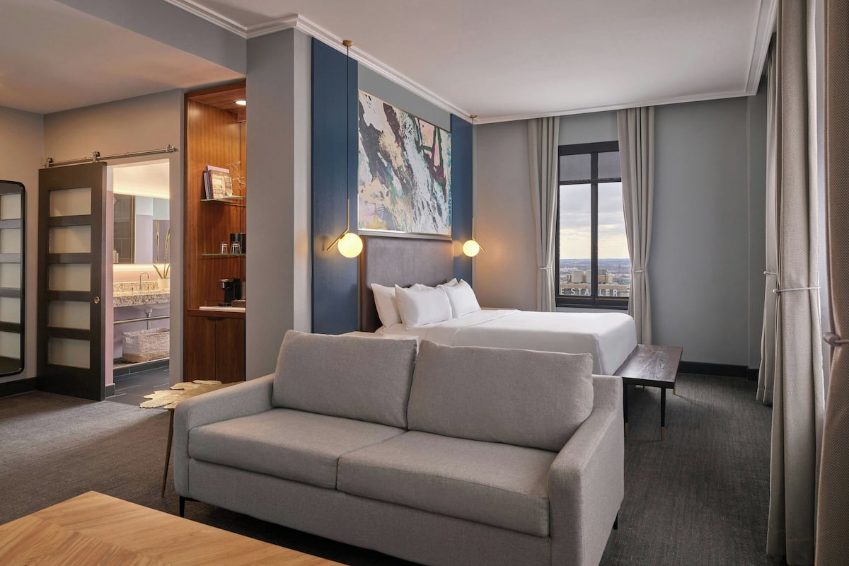A hotel room with a sofa, a bed, a desk, a bookshelf, and a large window.