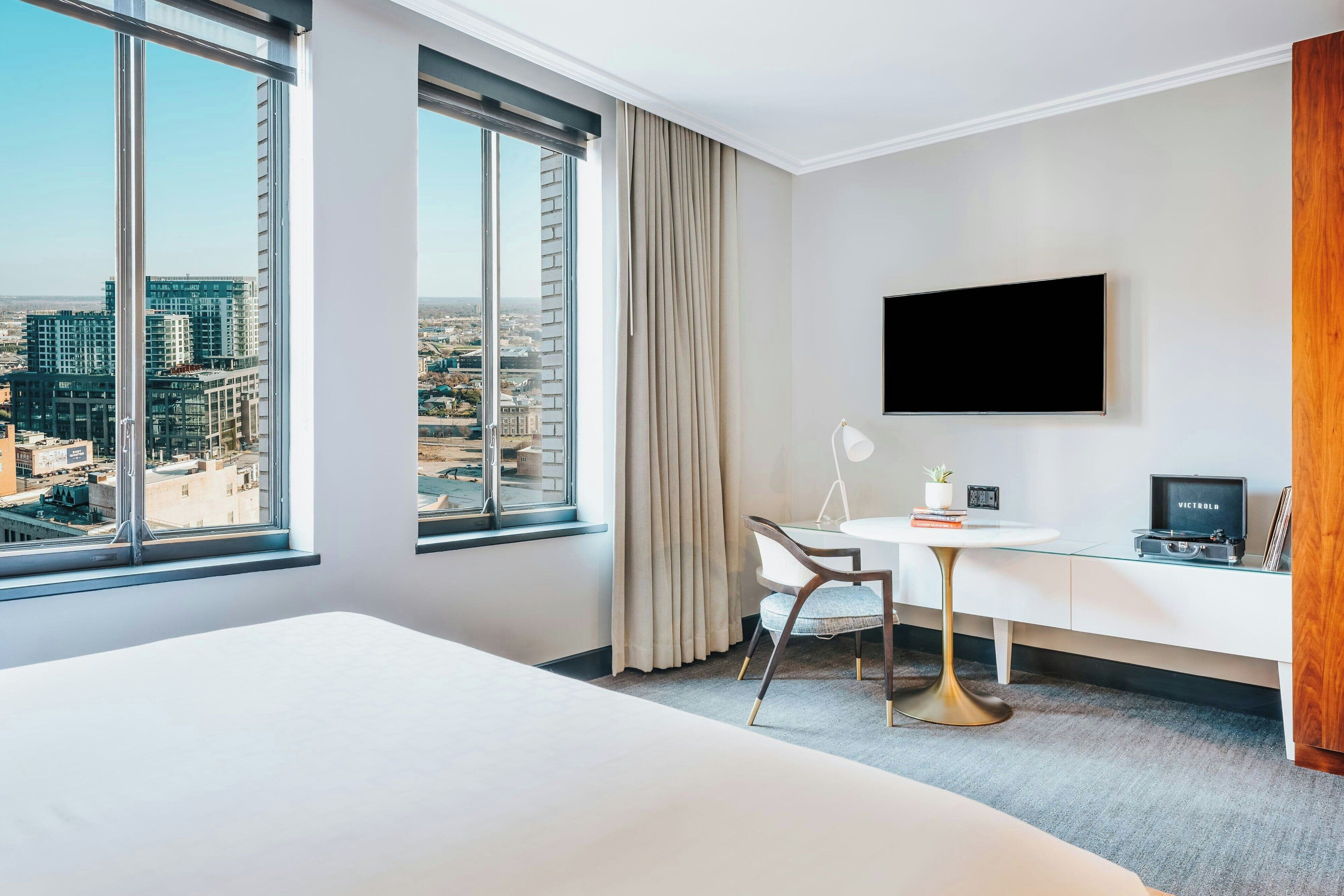 Habitación de hotel con una cama grande, un escritorio con una silla, un televisor montado en la pared y vistas de la ciudad a través de amplios ventanales.