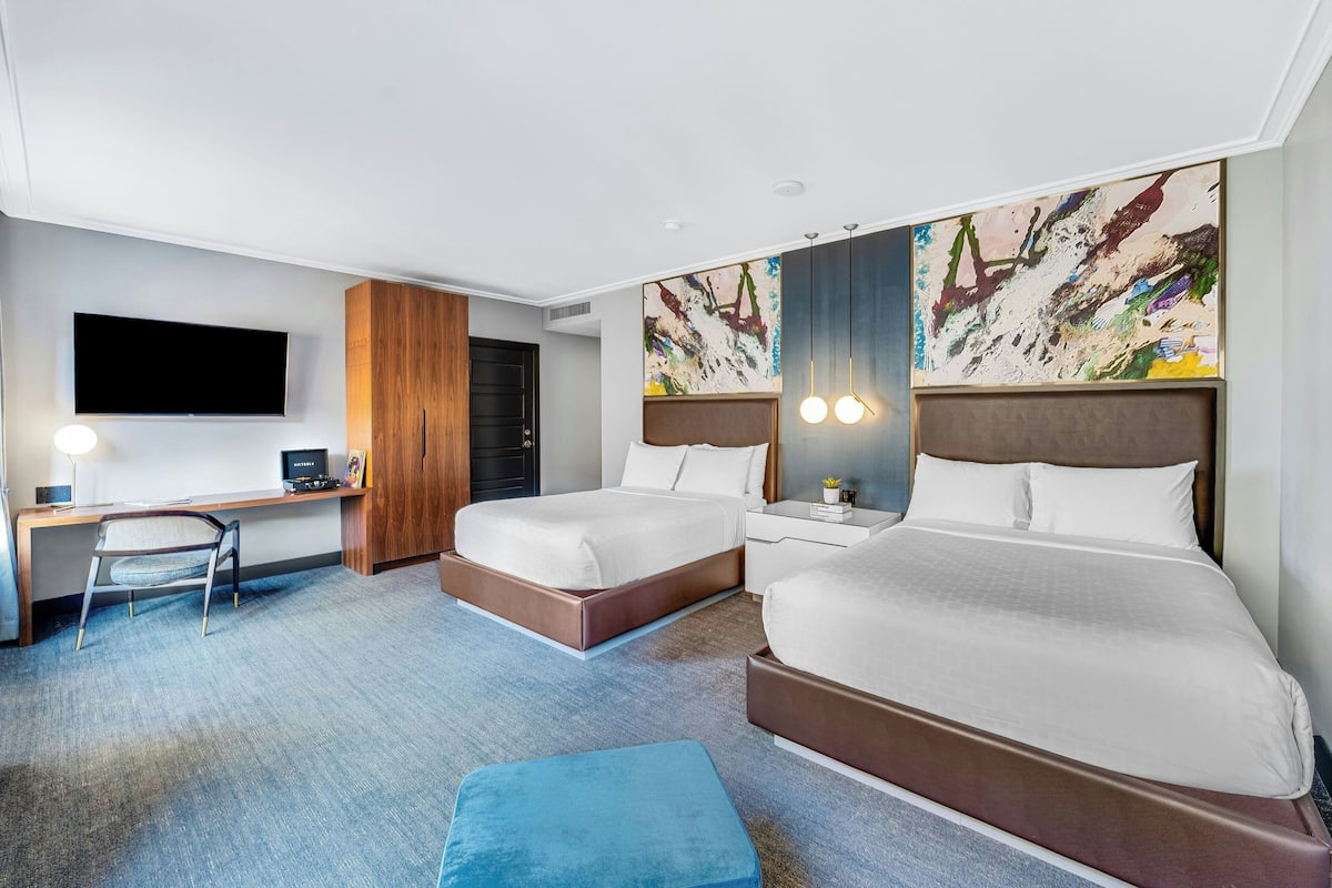 A hotel room with two beds, a desk with a chair, a flat-screen TV, and artwork on the walls.