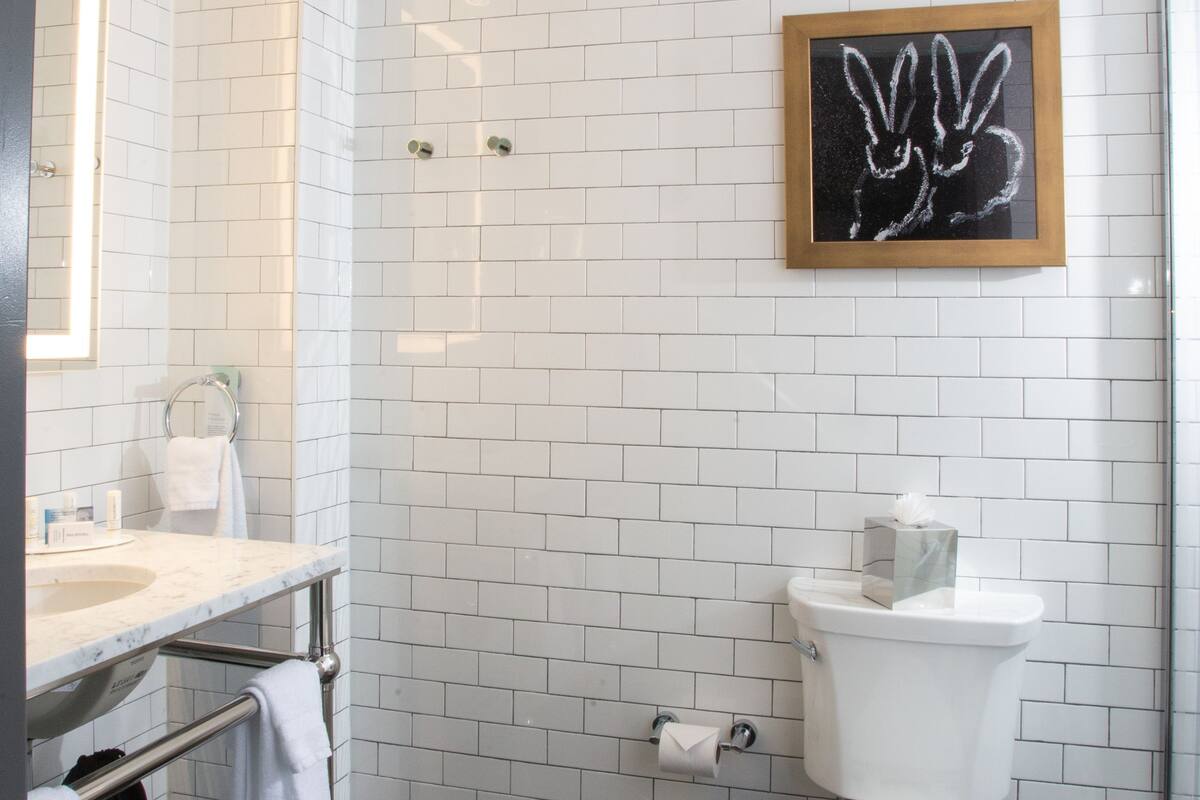 Ein modernes Badezimmer mit weißen Fliesen im U-Bahn-Stil, einer weißen Toilette, einem Edelstahlwaschbecken und einem gerahmten Kunstwerk an der Wand.