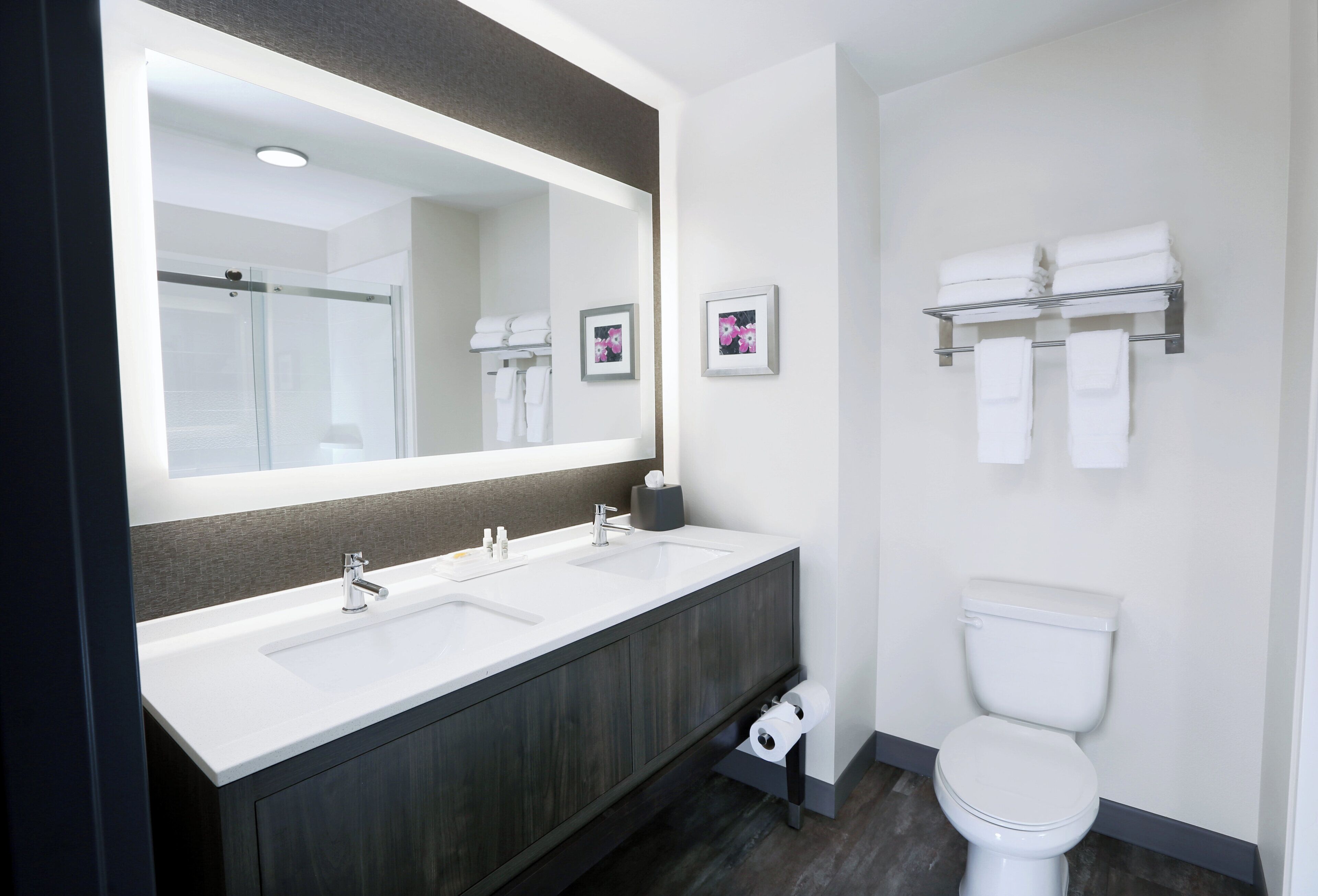 A modern bathroom with a large mirror, two sinks, a toilet, and a towel rack.