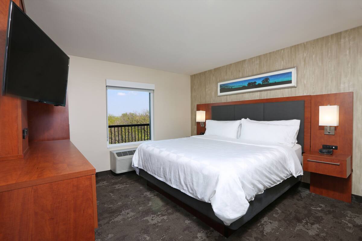 Une chambre d’hôtel avec un lit, un téléviseur fixé au mur, une fenêtre donnant sur un beau paysage et un tableau au-dessus du lit.