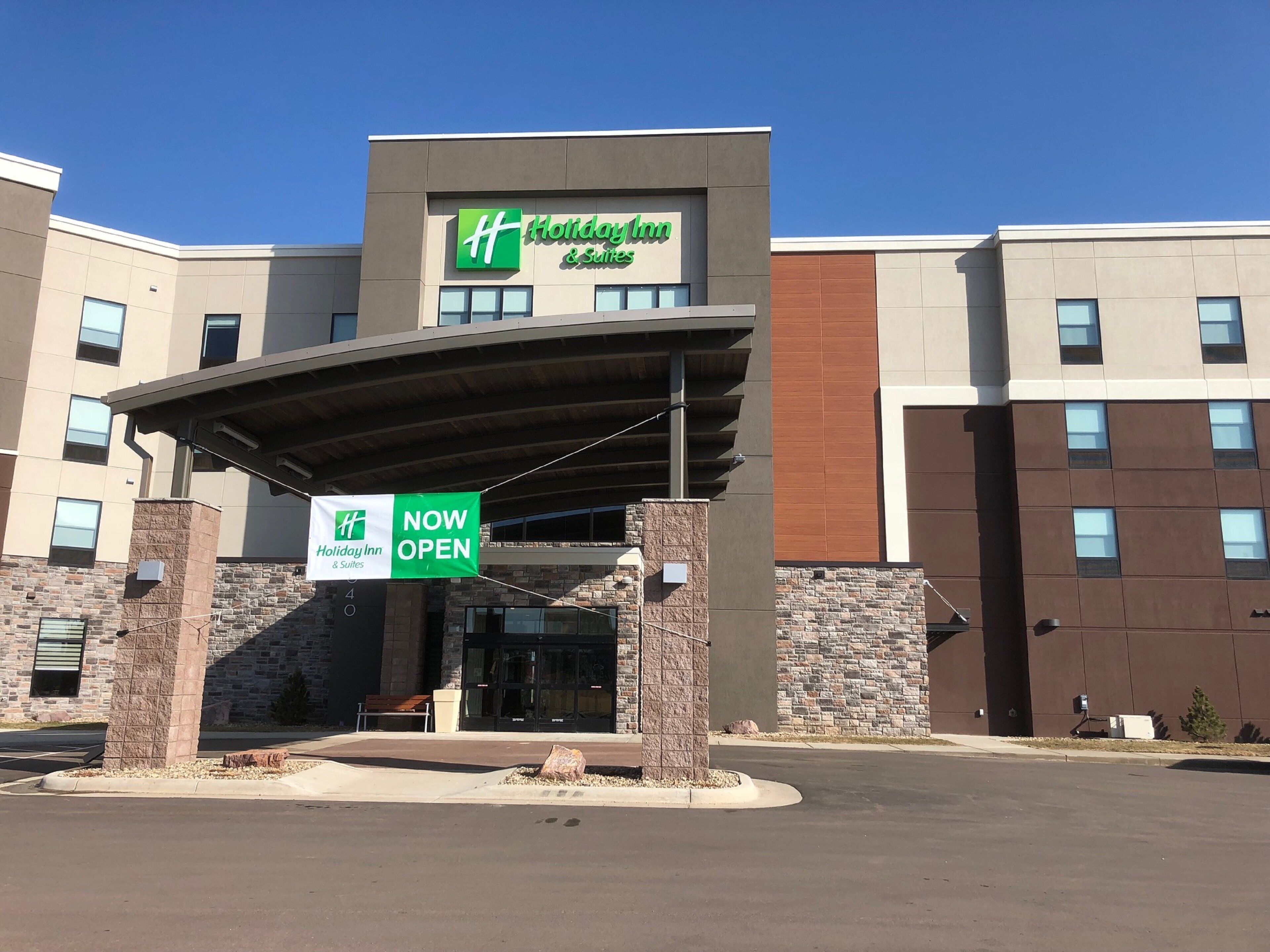 Edificio de Holiday Inn & Suites con un diseño arquitectónico moderno, destacando una entrada principal con un vestíbulo cubierto y señalización que indica que el establecimiento está ahora abierto.