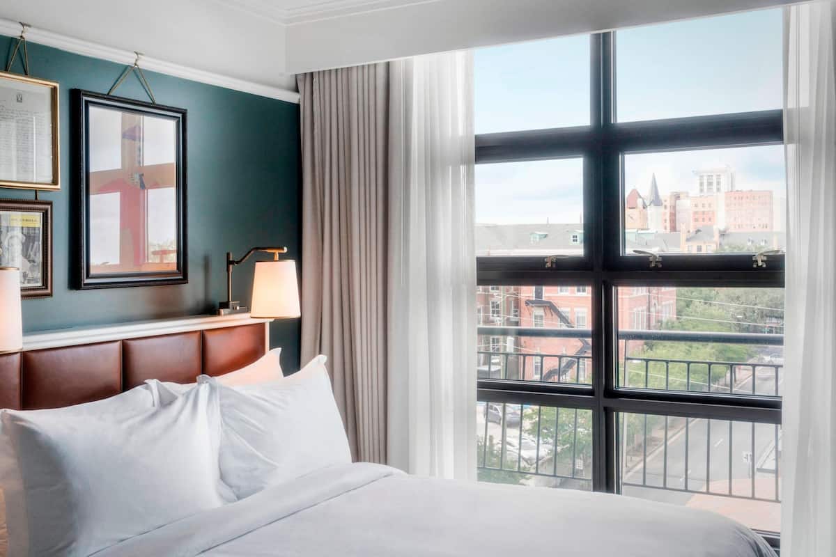 A hotel room with a large window, a bed with white linens, a headboard, and framed pictures on the wall.