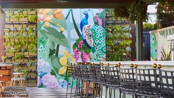 Uma área de bar com um mural de pavões e flores, bancos altos e chão xadrez.
