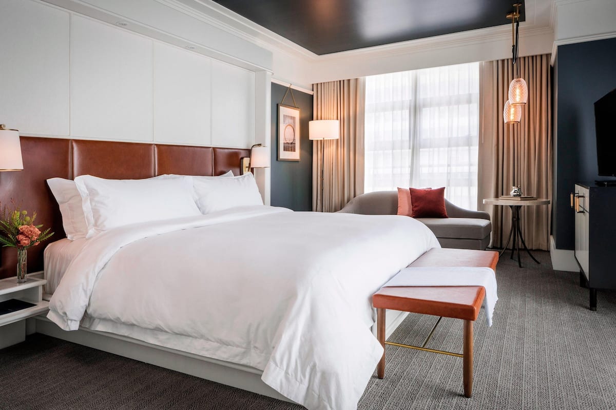 A hotel room with a large bed, a bedside table with a lamp, a small table with a lamp, a sofa, and a TV.