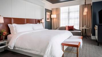 Quarto de hotel com uma cama grande, mesa de cabeceira com abajur, mesinha com luminária, um sofá e uma televisão.