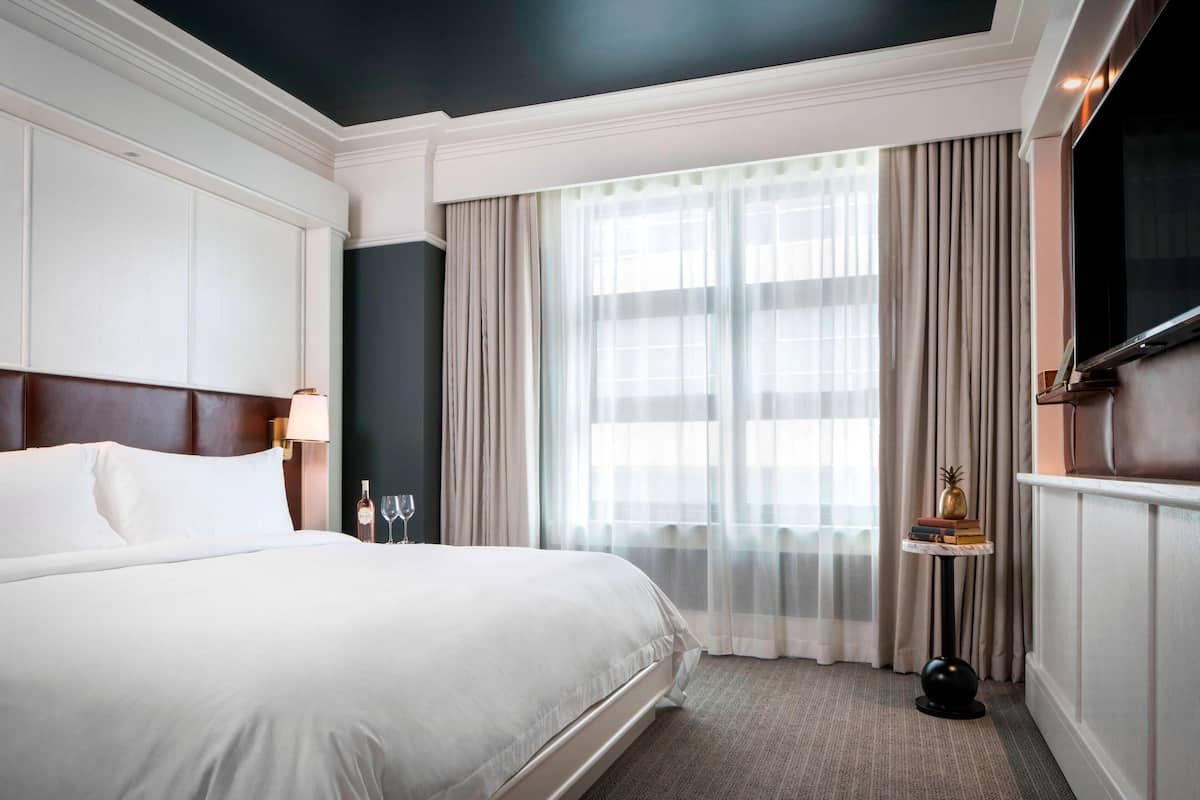 A hotel room with a large bed, a TV mounted on the wall, a small table with a lamp, and a window with sheer curtains.