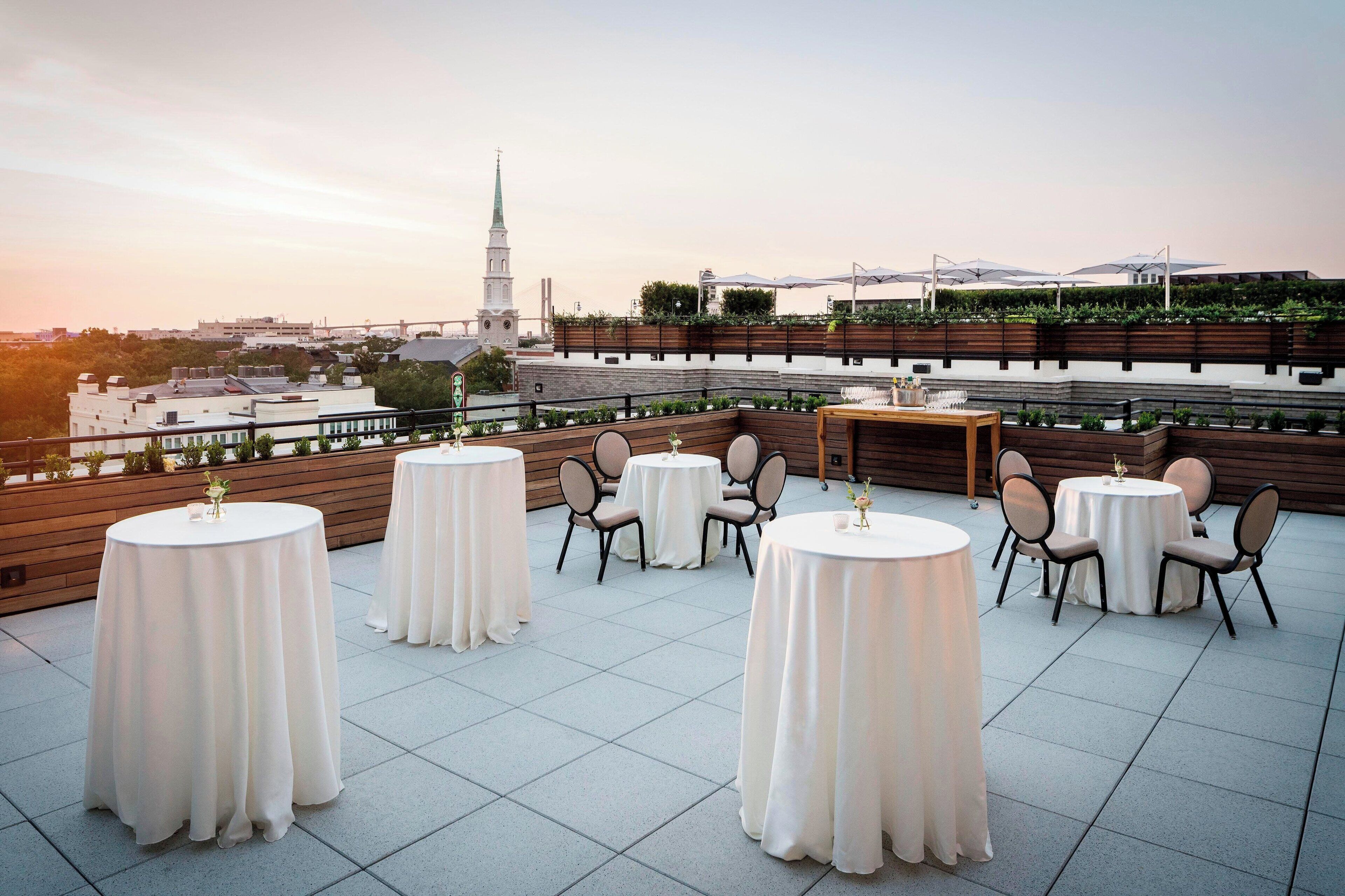 A rooftop terrace with round tables and chairs, a wooden deck, and a view of a cityscape including a church spire.
