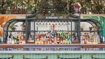 Een bar met verschillende drankflessen en glazen, een decoratief metalen hekwerk en potplanten.