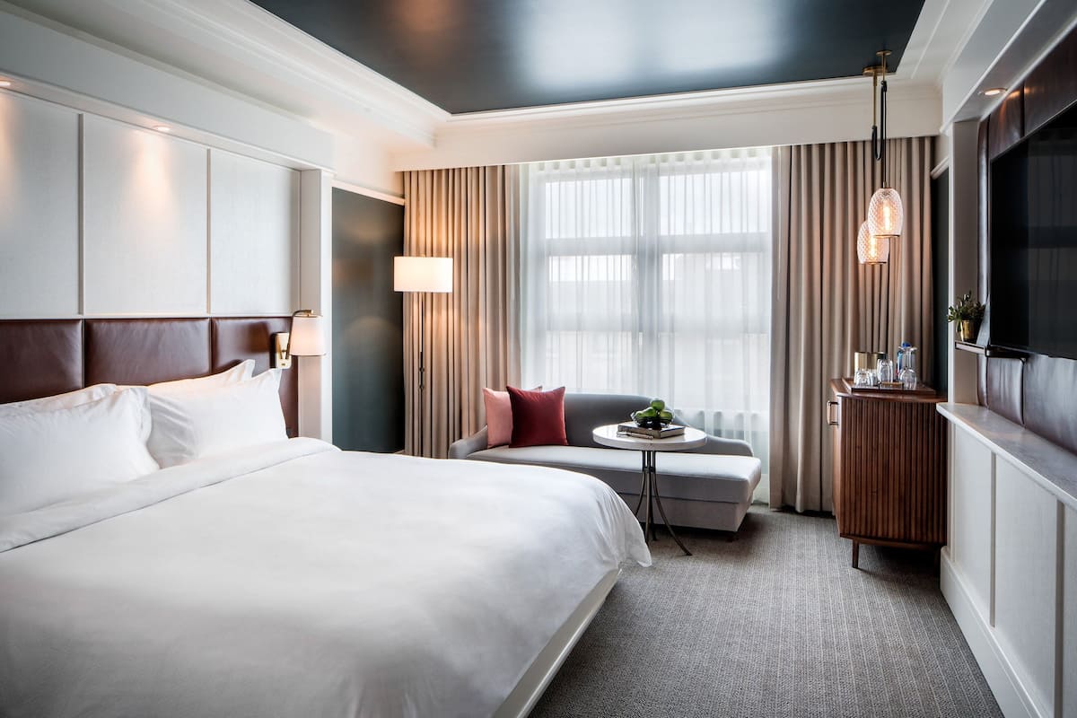 A hotel room with a large bed, a sofa, a small table with a plant, a TV, and a window with curtains.