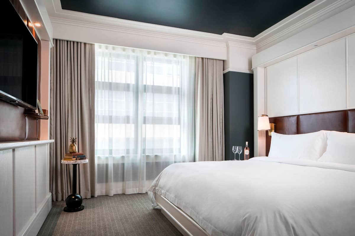 A hotel room with a large bed, a flat-screen TV, a small table with books and a pineapple, and a window with sheer curtains.